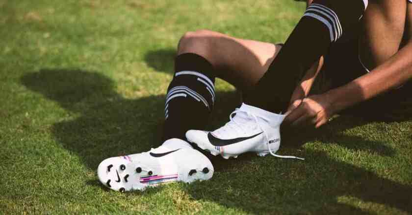 A football player sits on the grass, adjusting their white Nike cleats to ensure a proper fit, demonstrating how should football cleat fit, while wearing black socks with white stripes.